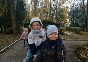 Chłopcy bawią się w ogródkowej lokomotywie, w tle idzie dziewczynka.