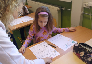 Dziewczynka siedzi w ławce i wykonuje zadanie na karcie pracy