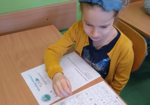 Dziewczynka siedzi w ławce i wykonuje zadanie na karcie pracy