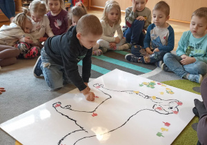 dzieci siedzą w kole. Na dywanie leży duża plansza psa
