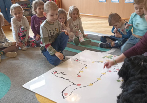 dzieci siedzą w kole. Na dywanie leży duża plansza psa