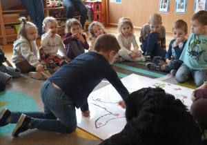 dzieci siedzą w kole. Na dywanie leży duża plansza psa