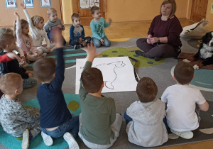 dzieci siedzą w kole. Na dywanie leży duża plansza psa