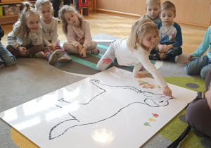 dzieci siedzą w kole. Na dywanie leży duża plansza psa