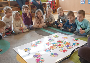 dzieci siedzą w kole. Na dywanie leży duża plansza psa