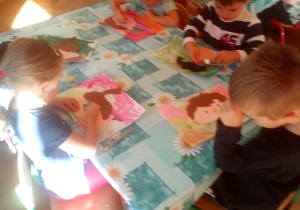 dzieci siedzą przy stole i naklejają kolorowe elementy z papieru