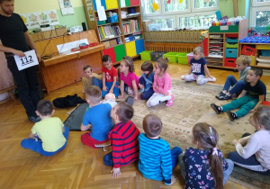 Dzieci siedzą na dywanie i słuchają strażaka mówiącego o numerach alarmowych