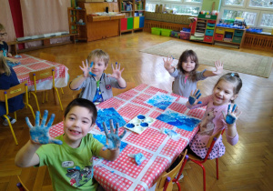 Dzieci siedzą przy stolikach, pokazują ubrudzone niebieska farba dłonie