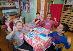 Dzieci siedzą przy stolikach, pokazują ubrudzone niebieska farba dłonie