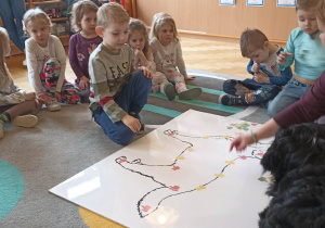 dzieci siedzą w kole. Na dywanie leży duża plansza psa