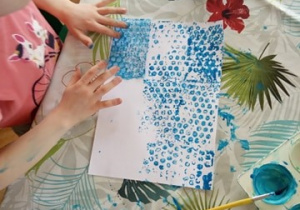 Dziewczynka odbija niebieską farbę na kartce papieru.
