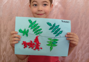 Chłopiec prezentuje swoją pracę - kontur gałązki jarzębiny wyklejony zielona plastelina i kulkami z czerwonej bibuły