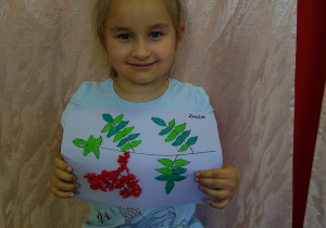 Dziewczynka prezentuje swoją pracę - kontur gałązki jarzębiny wyklejony zielona plastelina i kulkami z czerwonej bibuły