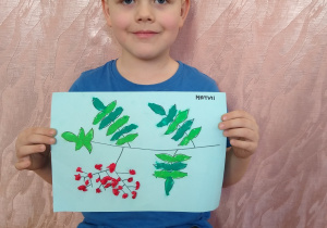 Chłopiec prezentuje swoją pracę - kontur gałązki jarzębiny wyklejony zielona plastelina i kulkami z czerwonej bibuły