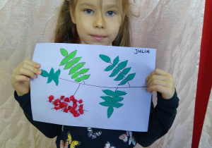 Dziewczynka prezentuje swoją pracę - kontur gałązki jarzębiny wyklejony zielona plastelina i kulkami z czerwonej bibuły