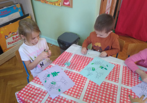 Dzieci wyklejają zielona plastelina i kulkami z czerwonej bibuły kontur gałązki jarzębiny