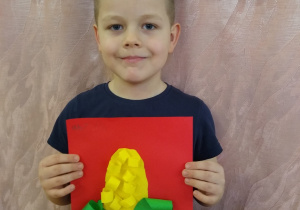 Chłopiec prezentuje swoją pracę - kukurydza z zielonego i żółtego papieru na czerwonym tle