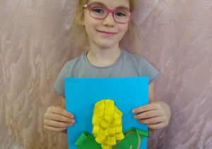 Dziewczynka prezentuje swoją pracę - kukurydza z zielonego i żółtego papieru na niebieskim tle