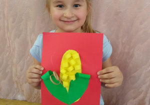 Dziewczynka prezentuje swoją pracę - kukurydza z zielonego i żółtego papieru na czerwonym tle