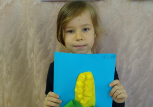 Dziewczynka prezentuje swoją pracę - kukurydza z zielonego i żółtego papieru na niebieskim tle