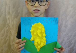 Chłopiec prezentuje swoją pracę - kukurydza z zielonego i żółtego papieru na niebieskim tle